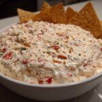 A white bowl filled with creamy, chunky boat dip speckled with red peppers and herbs, served with tortilla chips.