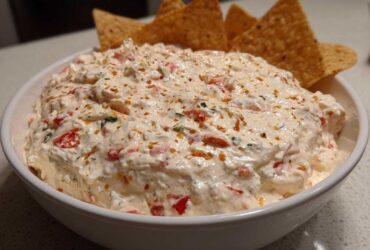 A white bowl filled with creamy, chunky boat dip speckled with red peppers and herbs, served with tortilla chips.
