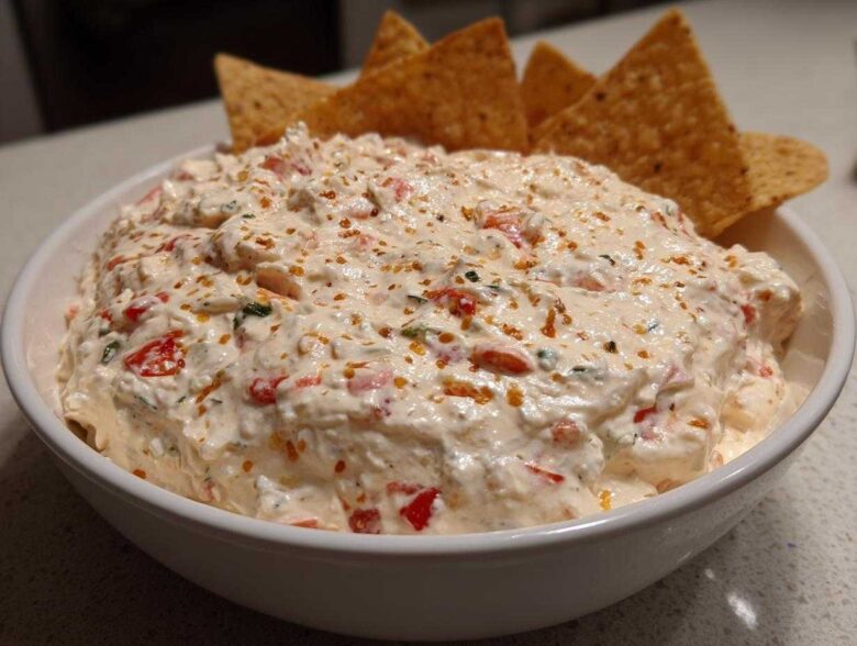 A white bowl filled with creamy, chunky boat dip speckled with red peppers and herbs, served with tortilla chips.