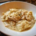 Close-up of a white bowl filled with creamy chicken and dumplings, topped with shredded chicken and herbs.