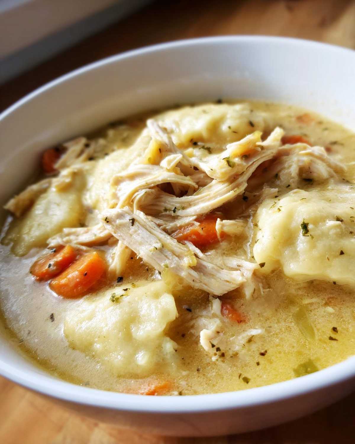 Close-up of a white bowl filled with creamy chicken and dumplings, featuring shredded chicken and sliced carrots.