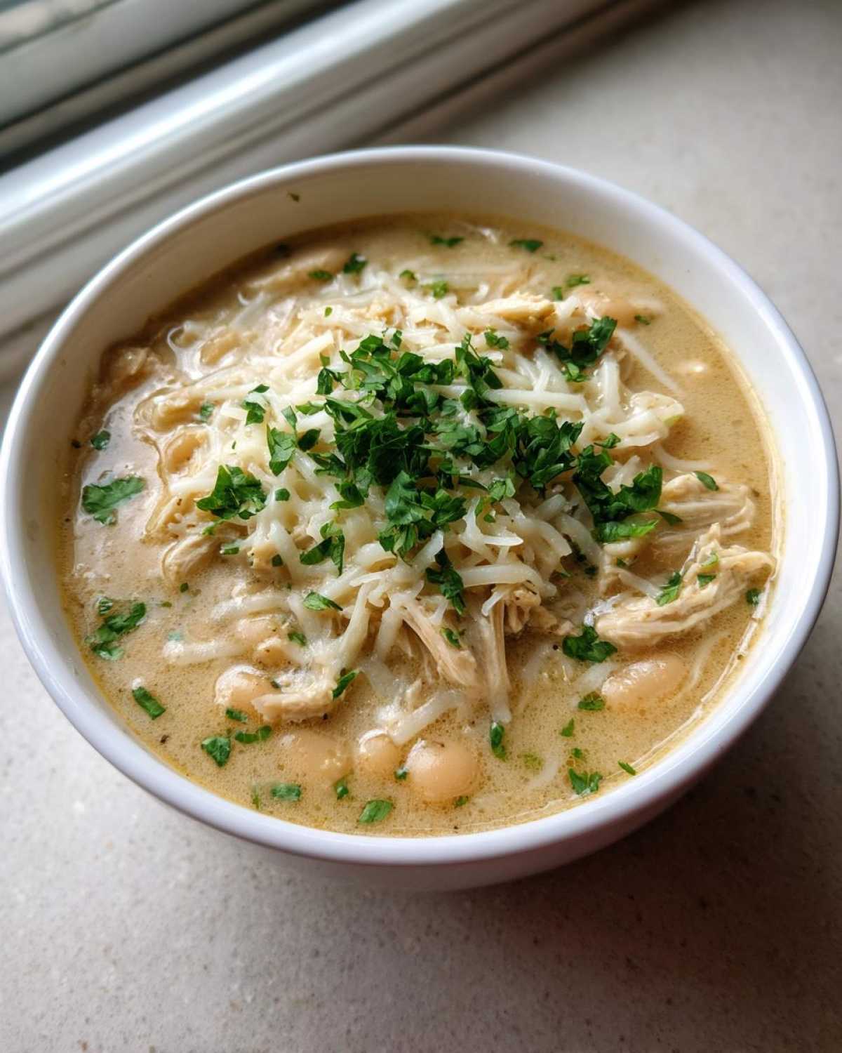 Close-up of a white bowl filled with creamy chicken chili, topped with shredded cheese and fresh parsley.