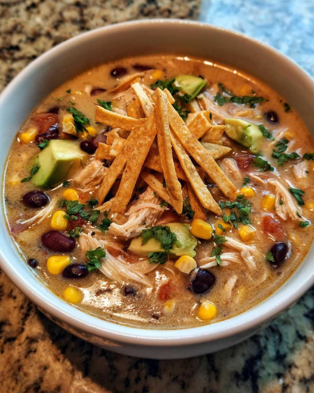 A close-up of creamy chicken tortilla soup, loaded with shredded chicken, black beans, corn, avocado, and crispy tortilla strips.