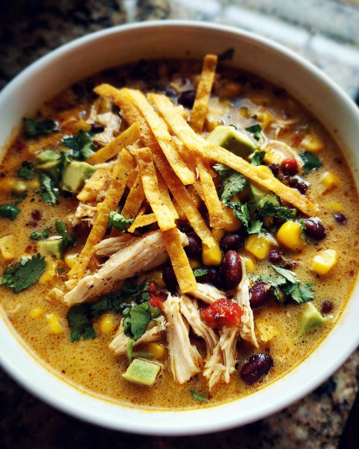 Close-up of a bowl of creamy chicken tortilla soup topped with shredded chicken, avocado, beans, and crispy tortilla strips.