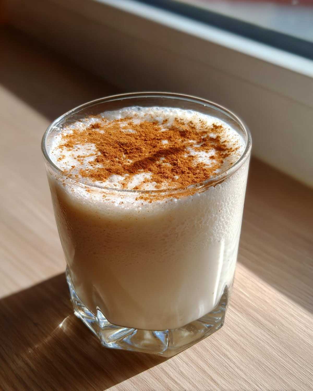 A glass of creamy, pale coquito topped generously with ground cinnamon, sitting on a wooden surface near a window.