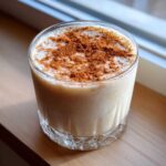 A glass of creamy, light brown coquito topped generously with ground cinnamon.