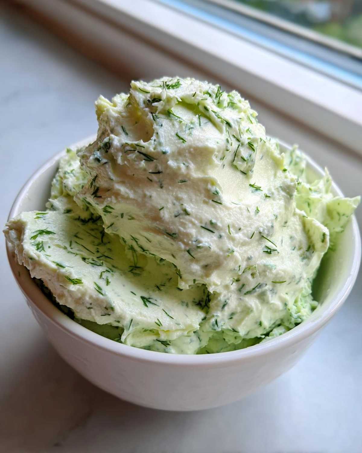 A close-up of fluffy, pale green dill dip generously mixed with fresh dill, served in a white bowl.