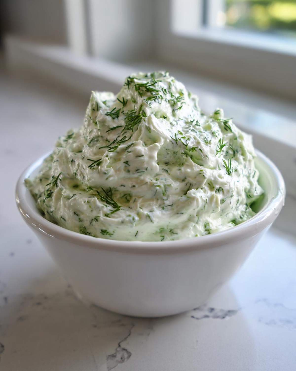 A white bowl filled with creamy, bright green dill dip, generously topped with fresh chopped dill.