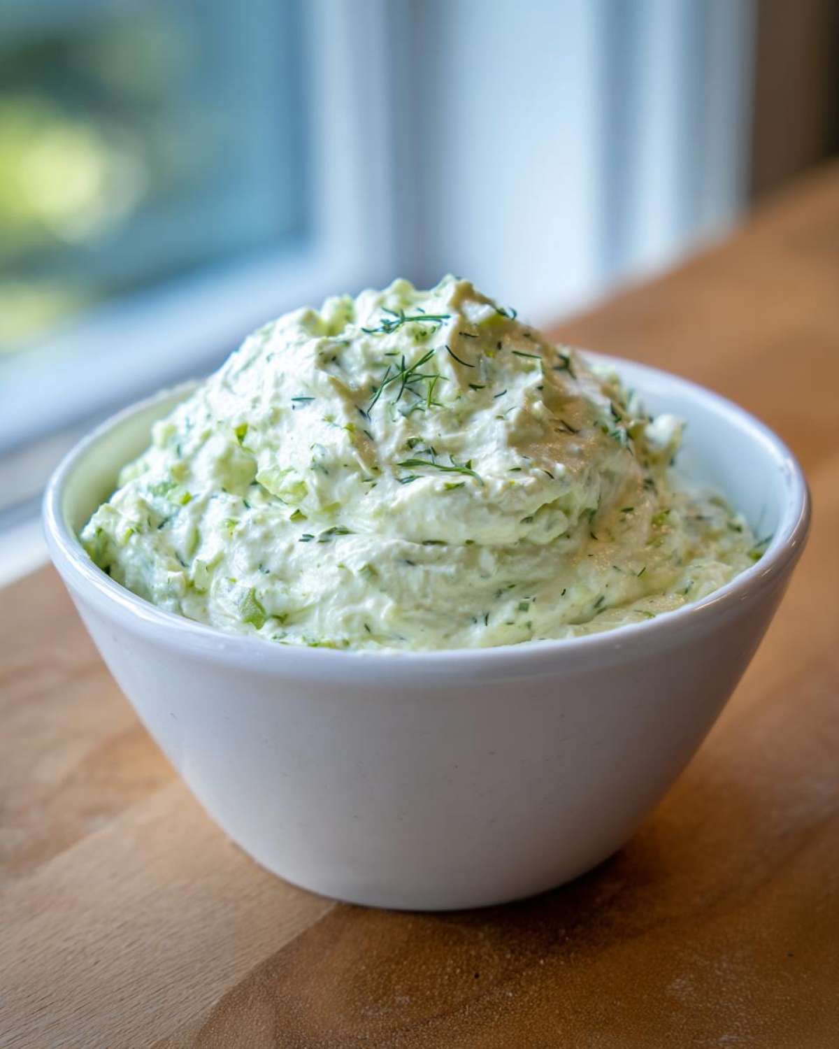 A close-up of creamy dill pickle dip, topped with fresh dill, served in a white bowl on a wooden surface.