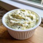 A close-up of creamy, thick dill pickle dip speckled with fresh dill, served in a white fluted ramekin.