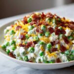 A close-up of creamy pea salad mixed with a white dressing, topped generously with crumbled bacon and shredded cheddar cheese.