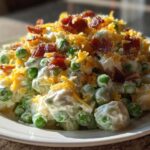 A close-up of creamy pea salad mixed with a white dressing, topped generously with crumbled bacon and shredded cheddar cheese.