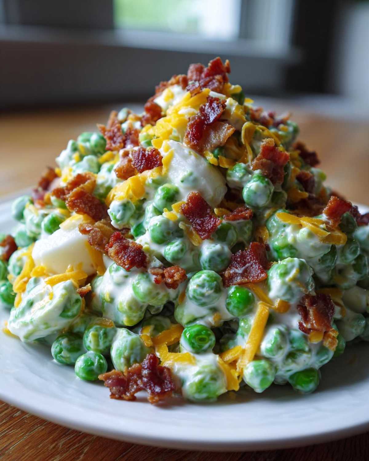 A mound of creamy pea salad mixed with dressing, topped generously with crumbled bacon and shredded cheddar cheese.
