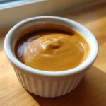 A close-up of thick, creamy peanut sauce served in a small, white, fluted ramekin on a wooden surface.