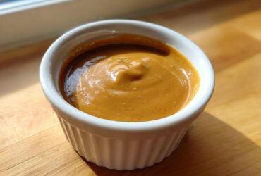 A close-up of thick, creamy peanut sauce served in a small, white, fluted ramekin on a wooden surface.