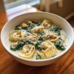 A white bowl filled with creamy tortellini soup, featuring cheese tortellini, wilted spinach, and topped with grated parmesan.