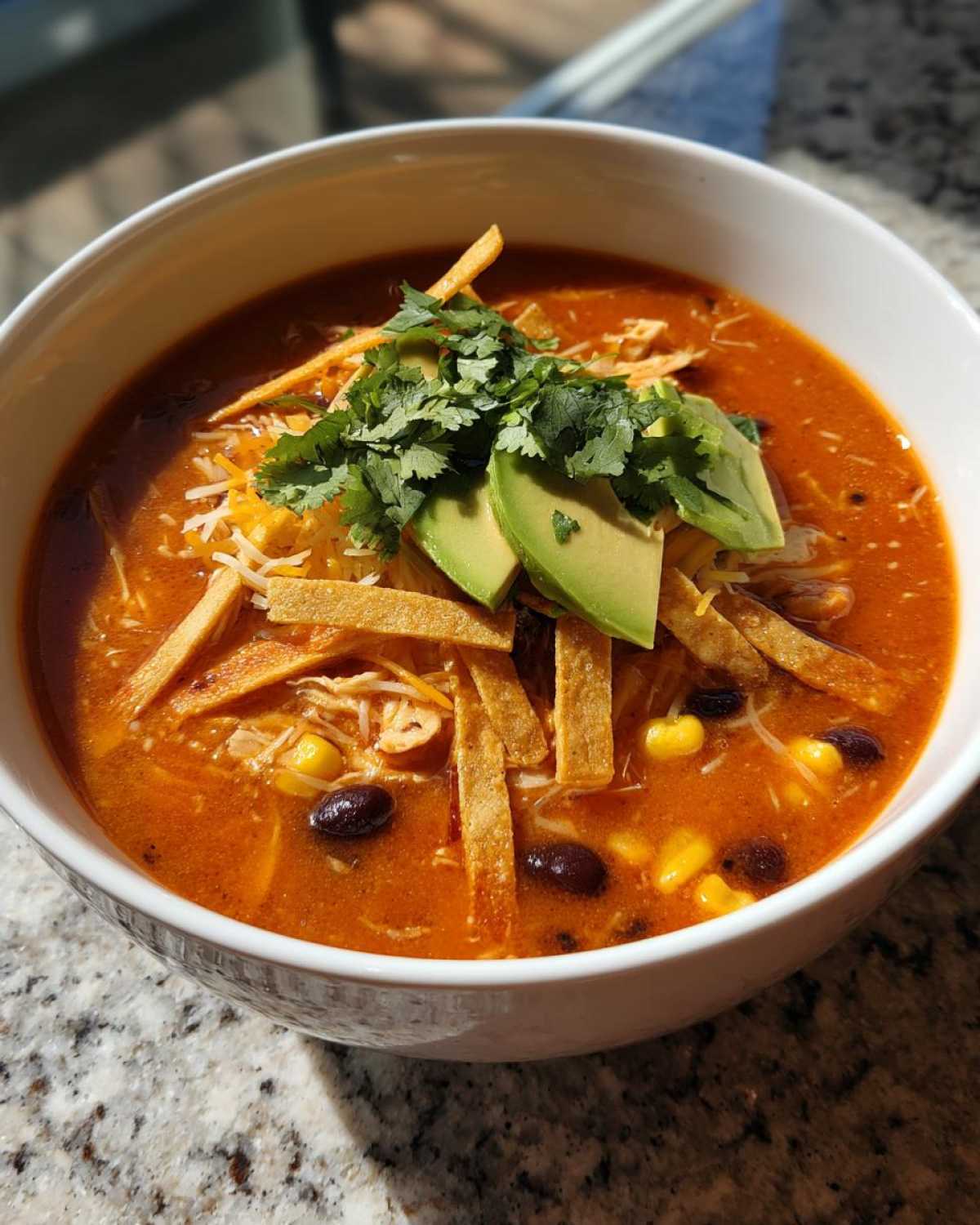 A close-up of a bowl of creamy tortilla soup recipe, topped with avocado slices, cilantro, cheese, and tortilla strips.