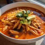 A close-up of a creamy tortilla soup recipe bowl filled with shredded chicken, black beans, corn, topped with tortilla strips, avocado, and cilantro.