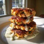 A stack of three crispy, golden-brown salmon cakes drizzled with creamy dill sauce, photographed in bright sunlight.