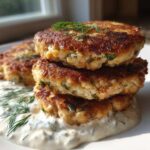 A stack of three golden-brown, crispy salmon cakes resting on a bed of creamy dill sauce, garnished with fresh dill.