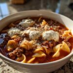 A close-up of a white bowl filled with rich, red crockpot lasagna soup, topped with dollops of ricotta and shredded Parmesan cheese.