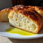 Close-up of a thick slice of crusty Italian bread showing an open, airy crumb dipped in bright green olive oil.