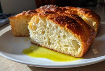 Close-up of a thick slice of crusty Italian bread showing an open, airy crumb dipped in bright green olive oil.