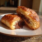 A golden, flaky chocolate croissant cut in half showing gooey, melted chocolate filling.