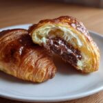 A flaky, golden brown chocolate croissant cut in half revealing a gooey, melted chocolate filling.