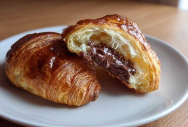 A flaky, golden brown chocolate croissant cut in half revealing a gooey, melted chocolate filling.