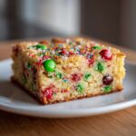 A close-up of a thick slice of christmas cookie bars studded with red and green M&Ms and colorful sprinkles.
