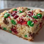 A thick, moist slice of christmas cookie bars topped with red and green M&Ms and festive sprinkles on a white plate.