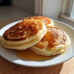 Three fluffy almond flour pancakes stacked on a white plate drizzled with maple syrup.
