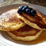A stack of three fluffy coconut flour pancakes drizzled with syrup and topped with fresh blueberries.