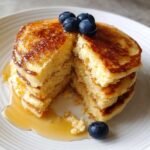 Close-up of a fluffy stack of coconut flour pancakes, cut open to show the texture, drizzled with syrup and topped with blueberries.