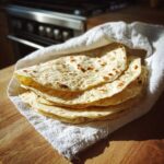 A stack of freshly cooked, warm corn tortillas wrapped in a white towel on a wooden surface.