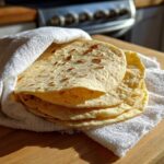 A stack of warm, freshly made corn tortillas wrapped in a white towel on a wooden surface.