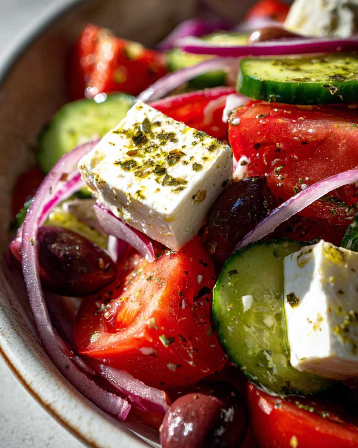 Close-up shot of a vibrant greek salad featuring large chunks of feta cheese seasoned with herbs, red tomatoes, cucumbers, and Kalamata olives.
