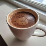 Close-up of a steaming white mug filled with rich, dark hot cocoa mix topped with small bubbles.