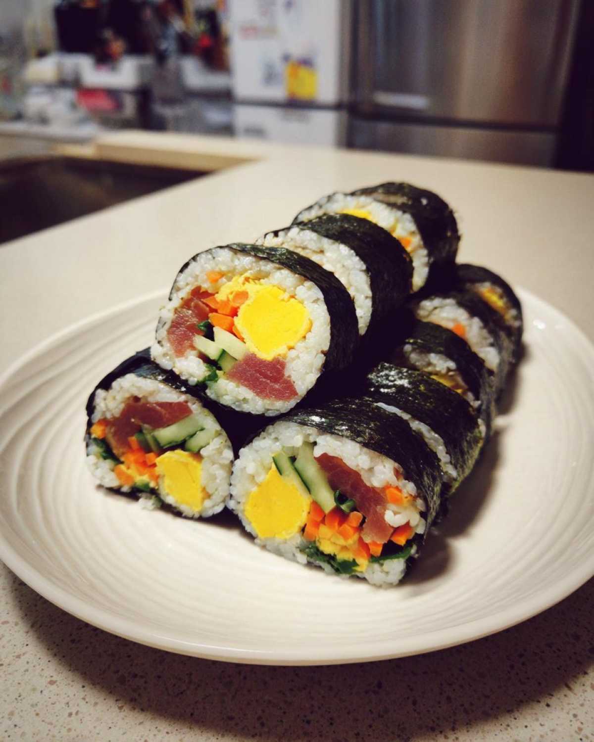 A stack of freshly sliced tuna kimbap rolls showing fillings like tuna, egg, cucumber, and carrot, resting on a white plate.