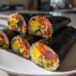 Close-up of several slices of homemade tuna kimbap featuring tuna, egg, carrots, and cucumber, beautifully arranged on a white plate.