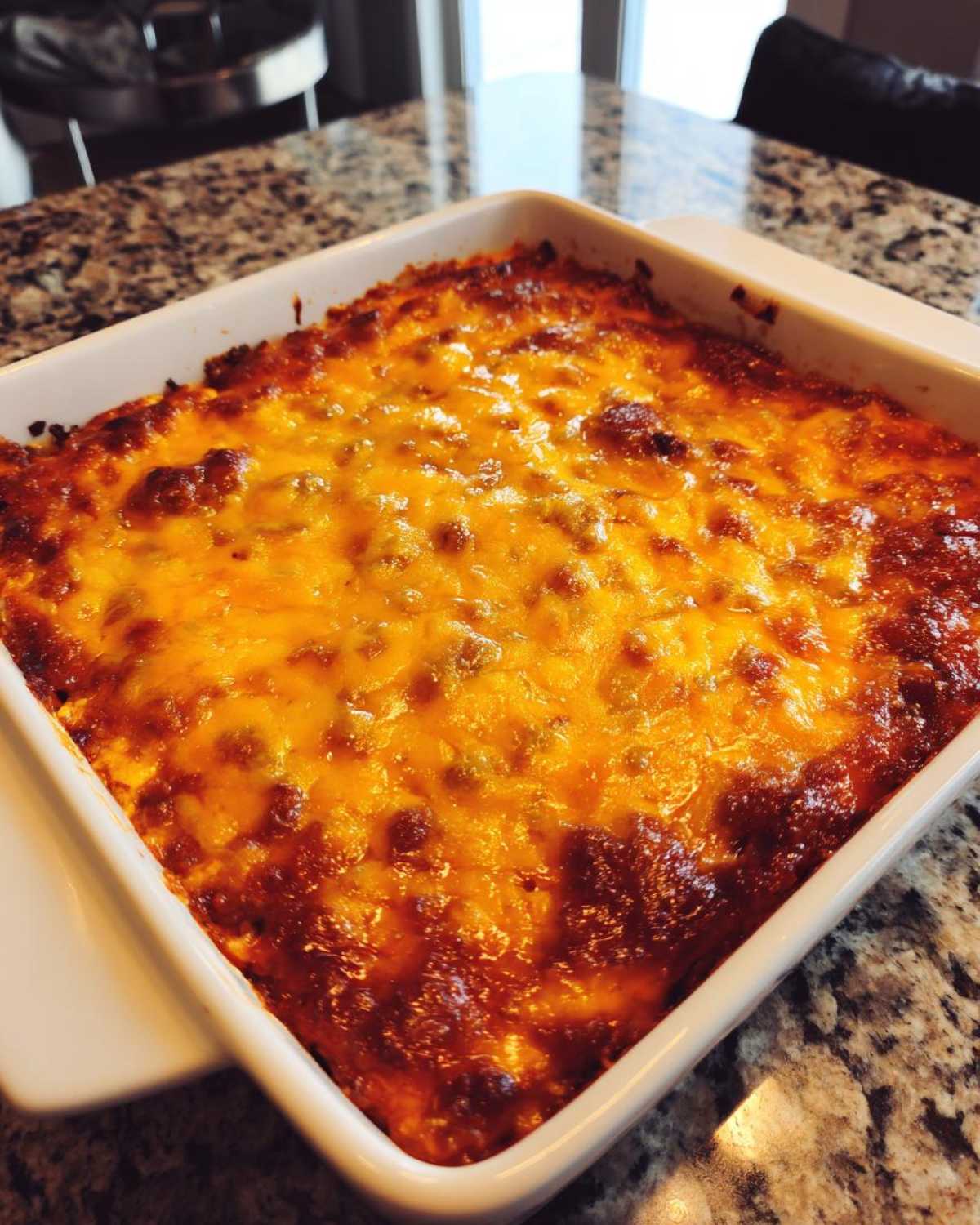 A square white baking dish filled with bubbling, baked chili cheese dip topped with melted, browned cheddar cheese.
