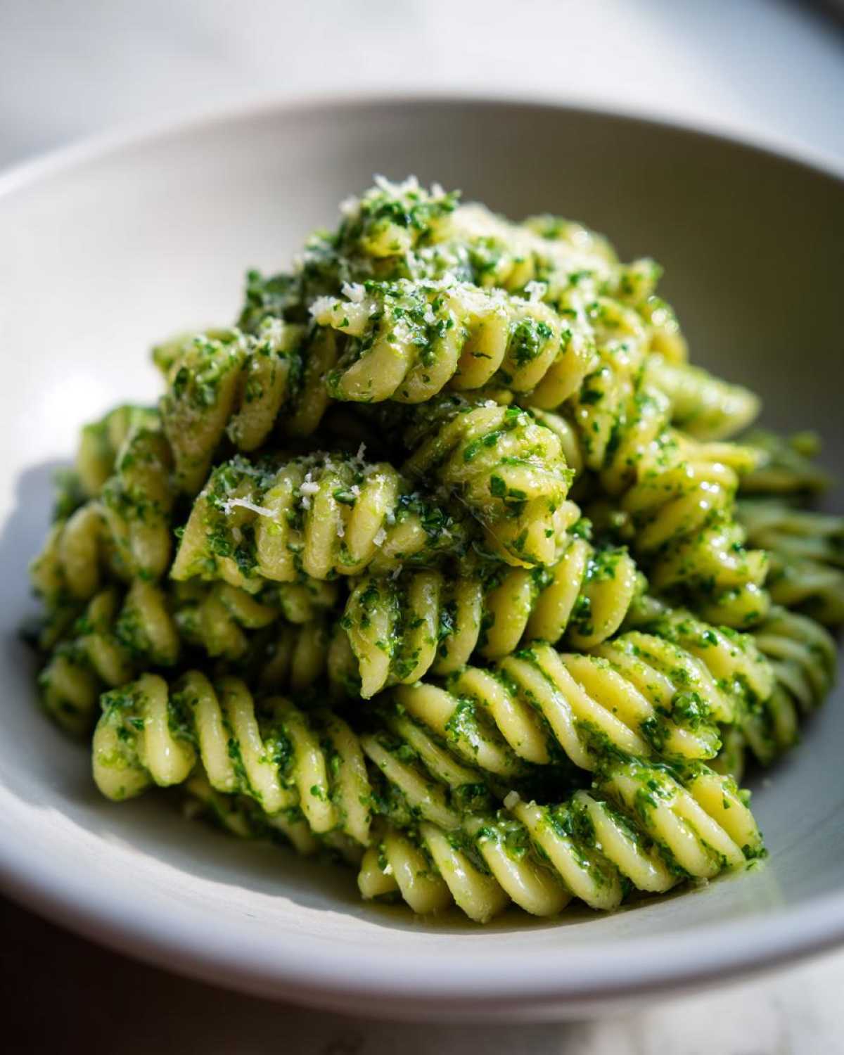 A close-up shot of fusilli pesto pasta tossed in vibrant green sauce and topped with grated Parmesan cheese.