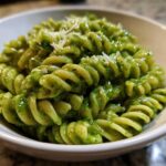 A close-up shot of vibrant green pesto pasta, made with fusilli noodles, topped with grated Parmesan cheese.