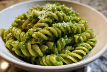 A close-up shot of vibrant green pesto pasta, made with fusilli noodles, topped with grated Parmesan cheese.