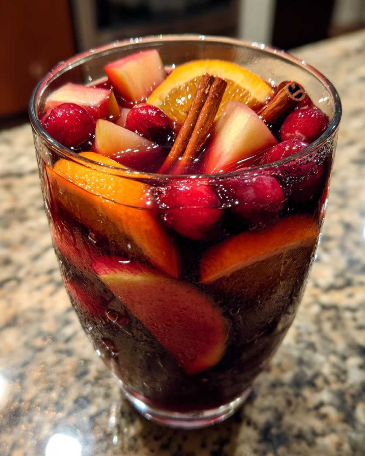 Close-up of a glass filled with cold christmas sangria, featuring orange slices, cranberries, apples, and cinnamon sticks.