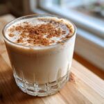 A glass of creamy coquito, topped generously with ground cinnamon, sitting on a wooden surface.