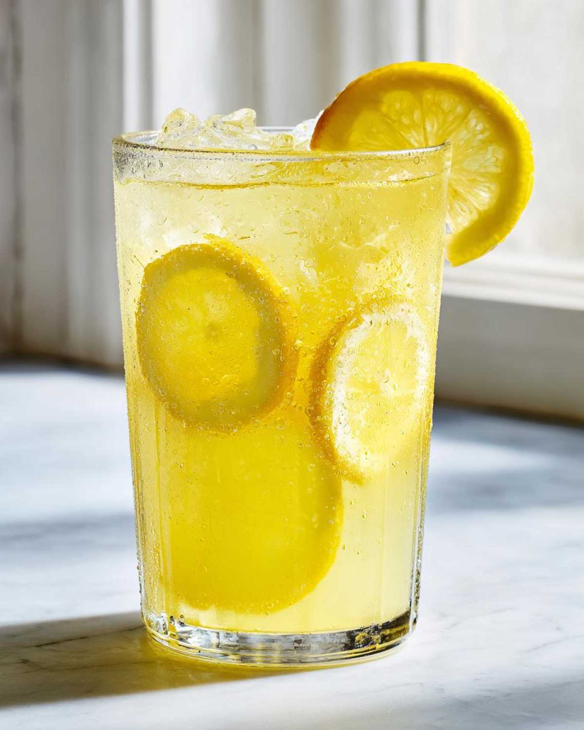 Close-up of a tall, sweating glass filled with bright yellow lemonade, ice, and lemon slices.