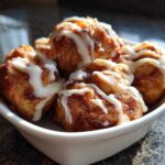 A white bowl filled with freshly baked, golden brown cinnamon roll bites drizzled generously with white icing.