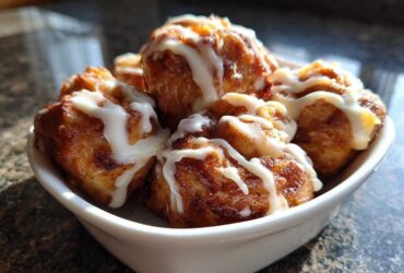 A white bowl filled with freshly baked, golden brown cinnamon roll bites drizzled generously with white icing.